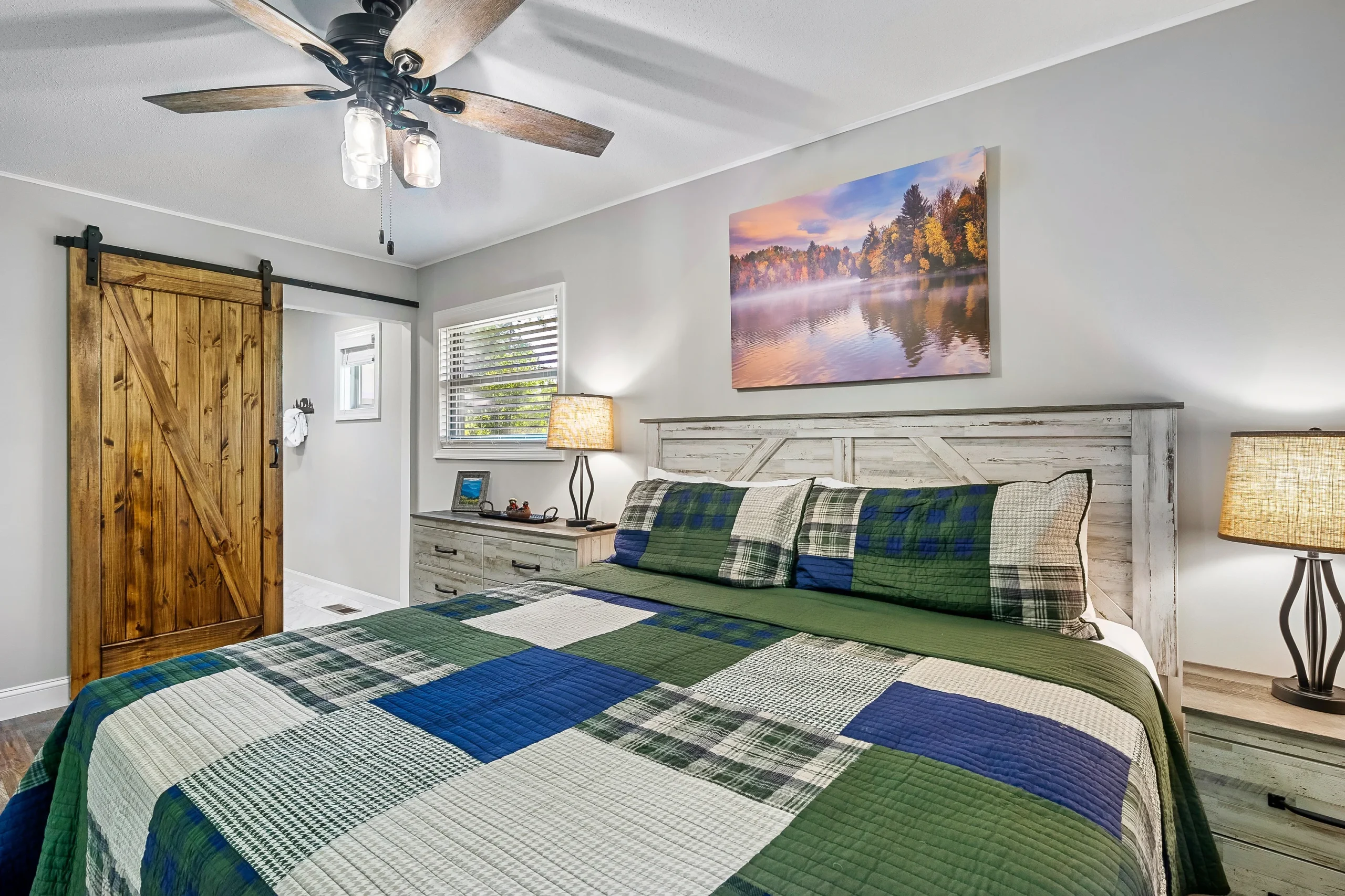 Farmhouse bedroom with whitewashed bed and navy plaid bedding, barn door closet, and wood-blade ceiling fan in a Pigeon