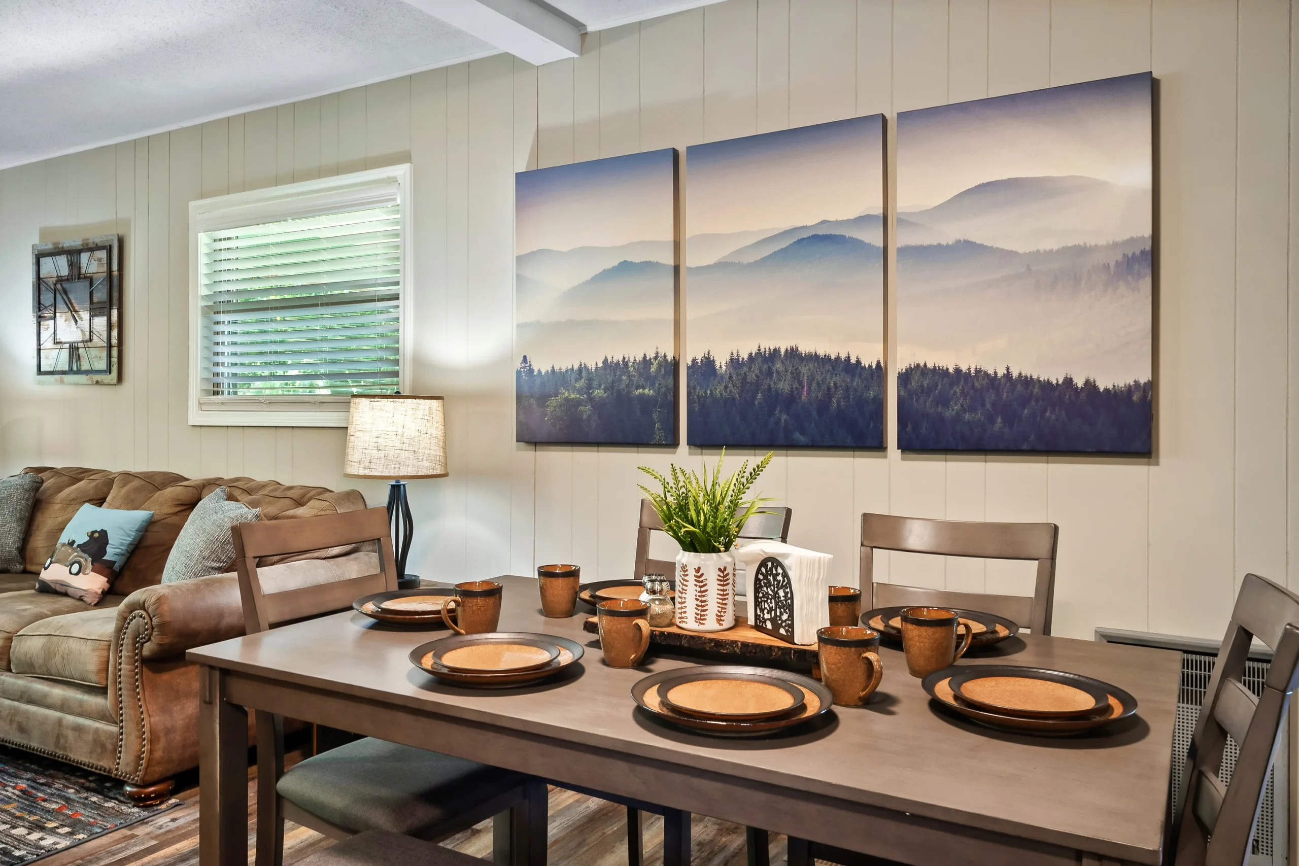 Open-concept dining room with wooden table set for four, cognac leather sofa, shiplap walls, and mountain triptych art at a
