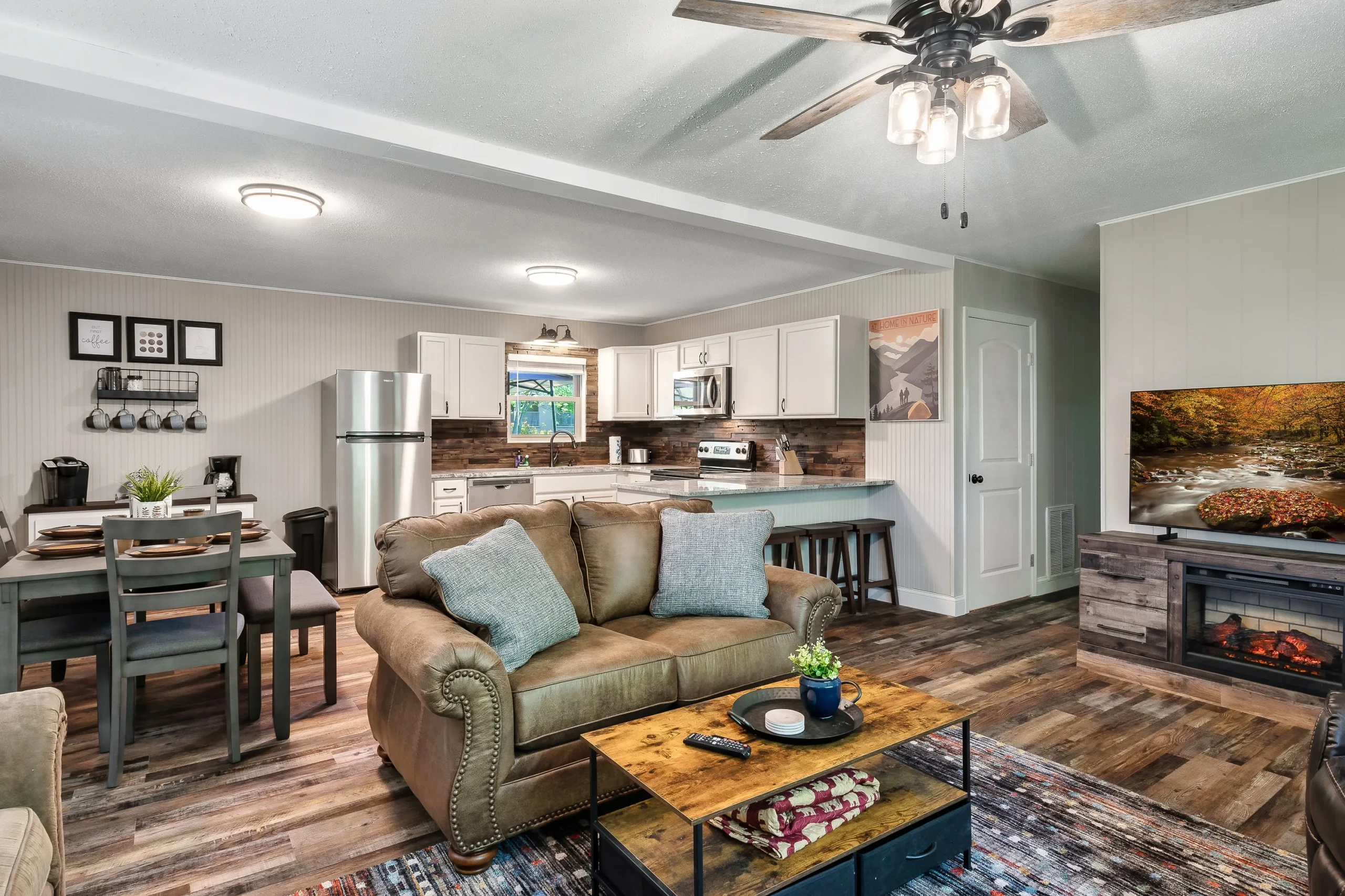 Open-concept living room with tan leather sofa, blue pillows, fireplace, and kitchen with white cabinets in a Pigeon Forge